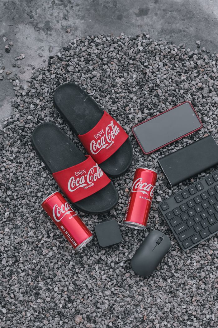 An artistic arrangement featuring Coca-Cola cans, sliders, a smartphone, mouse, and keyboard on gravel.
